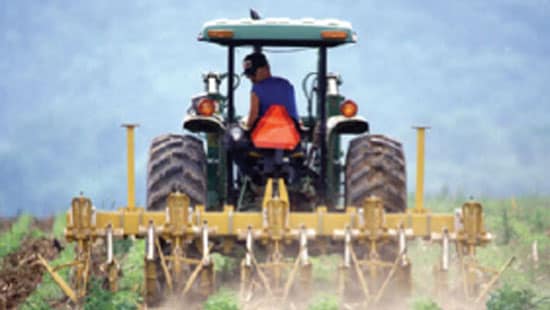 A man wearing sunglasses and baseball cap dragging a hoe line with a tractor on an open field.